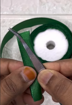 Two hands hold green ribbon with tweezers, folding it. In the background, a roll of green ribbon with a white center rests on a white tiled surface.
