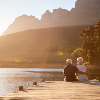An elderly couple sits on a wooden dock, embracing as they look out over a lake with mountains and trees in the background, bathed in warm sunset light.
