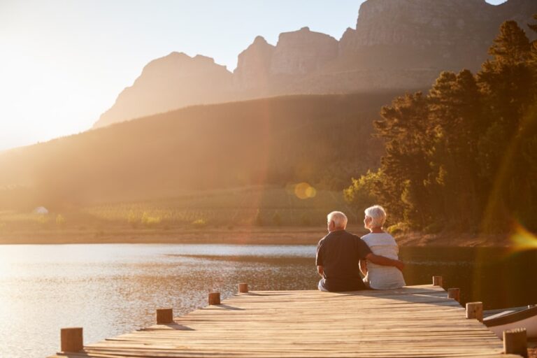 An elderly couple sits on a wooden dock, embracing as they look out over a lake with mountains and trees in the background, bathed in warm sunset light.