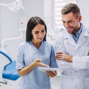 Two dental professionals in lab coats are standing in a bright dental office, smiling and discussing information displayed on a digital tablet. A dental chair and equipment are visible in the background.