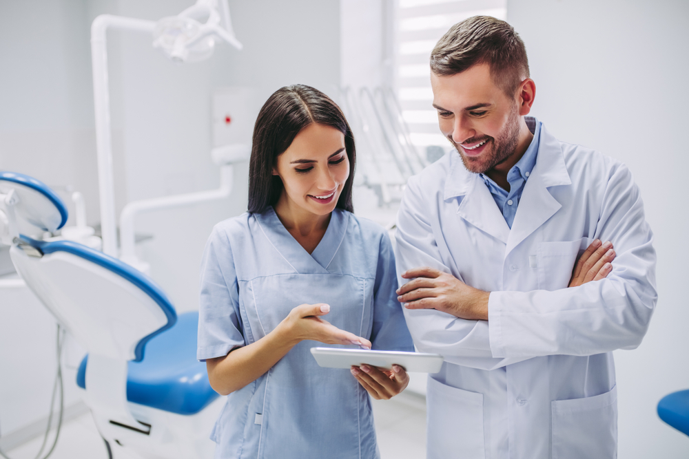 Two dental professionals in lab coats are standing in a bright dental office, smiling and discussing information displayed on a digital tablet. A dental chair and equipment are visible in the background.