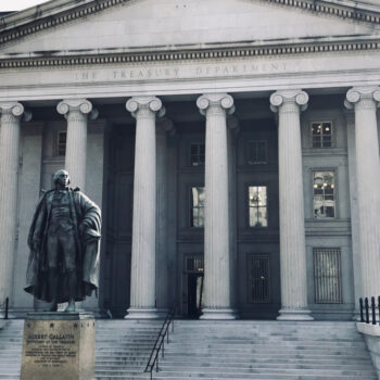 A neoclassical building with tall columns and The Treasury Department inscribed above the entrance; a statue of Albert Gallatin stands prominently in front.