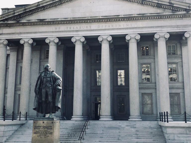 A neoclassical building with tall columns and The Treasury Department inscribed above the entrance; a statue of Albert Gallatin stands prominently in front.