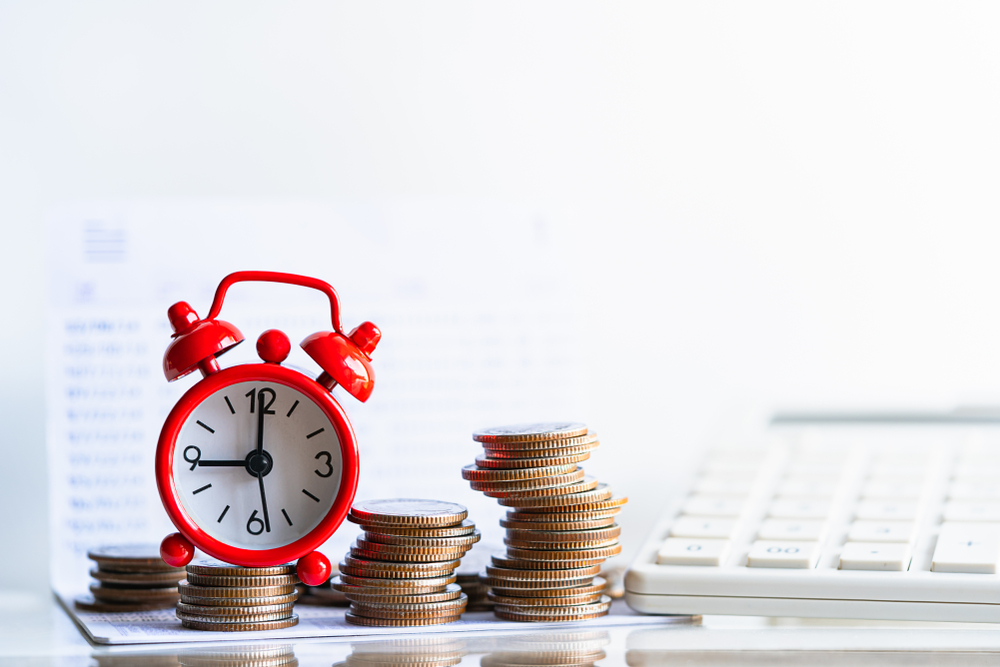 A small red alarm clock is placed among stacks of coins with a white calculator in the background, symbolizing the concepts of time, money, and financial planning.