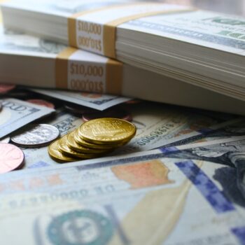 Stacks of $100 bills wrapped in $10,000 bands, gold coins, silver coins, and pennies scattered on top of more U.S. currency. The scene suggests wealth or financial abundance.