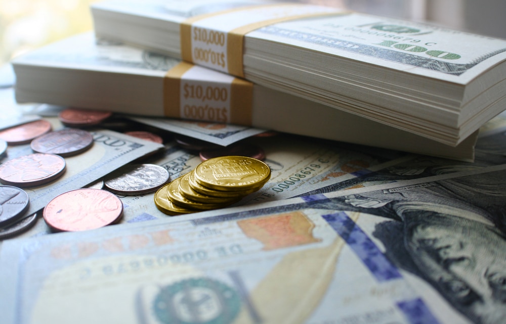 Stacks of $100 bills wrapped in $10,000 bands, gold coins, silver coins, and pennies scattered on top of more U.S. currency. The scene suggests wealth or financial abundance.