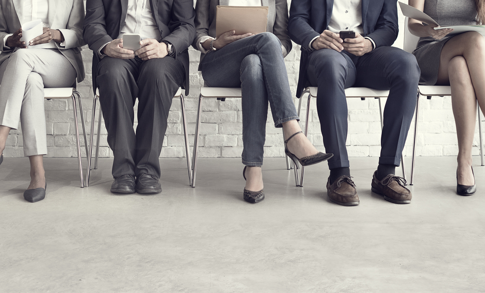 Five people sit in a row on chairs against a white brick wall, dressed in business attire, holding papers, tablets, or phones, with only their lower bodies and legs visible.