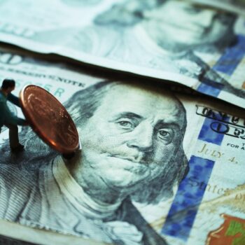 A tiny figurine pushes a large penny on top of two overlapping 100-dollar bills, highlighting the concept of financial struggle or value.