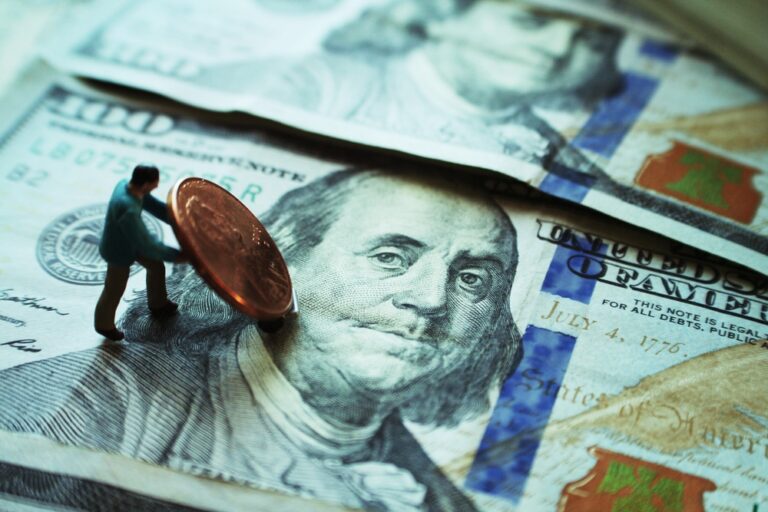 A tiny figurine pushes a large penny on top of two overlapping 100-dollar bills, highlighting the concept of financial struggle or value.
