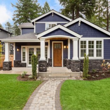 A charming blue house with white trim, a wooden front door, and a covered porch sits amid lush green grass and landscaped plants, with a stone path leading to the entrance. Tall trees are visible in the background.