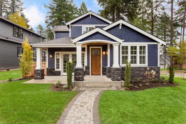 A charming blue house with white trim, a wooden front door, and a covered porch sits amid lush green grass and landscaped plants, with a stone path leading to the entrance. Tall trees are visible in the background.