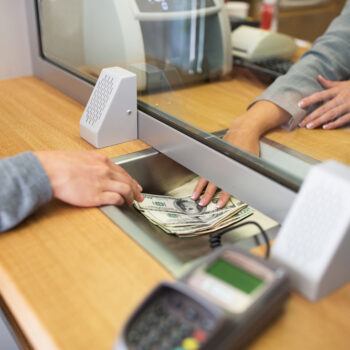 A person hands over a stack of U.S. dollar bills to a bank teller through a glass partition at a service counter. Both individuals’ hands are visible, along with speakers and a card reader on the counter.