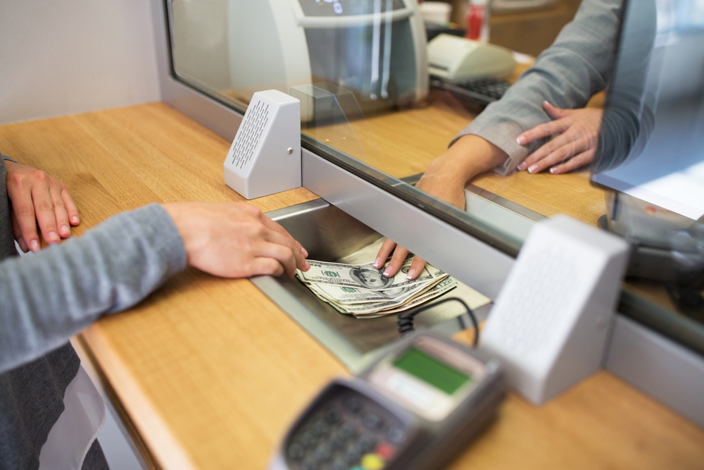 A person hands over a stack of U.S. dollar bills to a bank teller through a glass partition at a service counter. Both individuals’ hands are visible, along with speakers and a card reader on the counter.