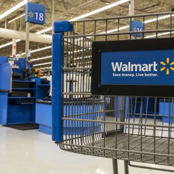 A Walmart shopping cart in the foreground with self-checkout registers and blue signs numbered 17 and 18 visible in the background inside a brightly lit Walmart store.