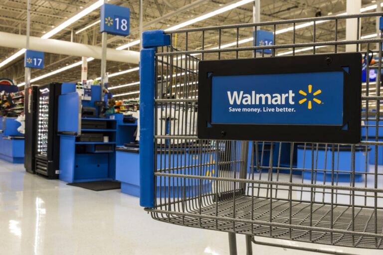 A Walmart shopping cart in the foreground with self-checkout registers and blue signs numbered 17 and 18 visible in the background inside a brightly lit Walmart store.