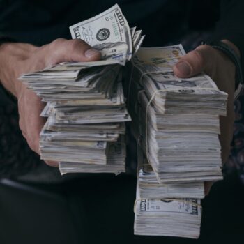 A person holding two large stacks of U.S. hundred-dollar bills, gripping the money tightly with both hands. The background and the persons clothing are dark and out of focus.
