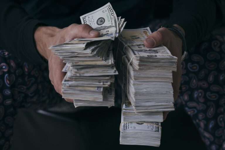 A person holding two large stacks of U.S. hundred-dollar bills, gripping the money tightly with both hands. The background and the persons clothing are dark and out of focus.