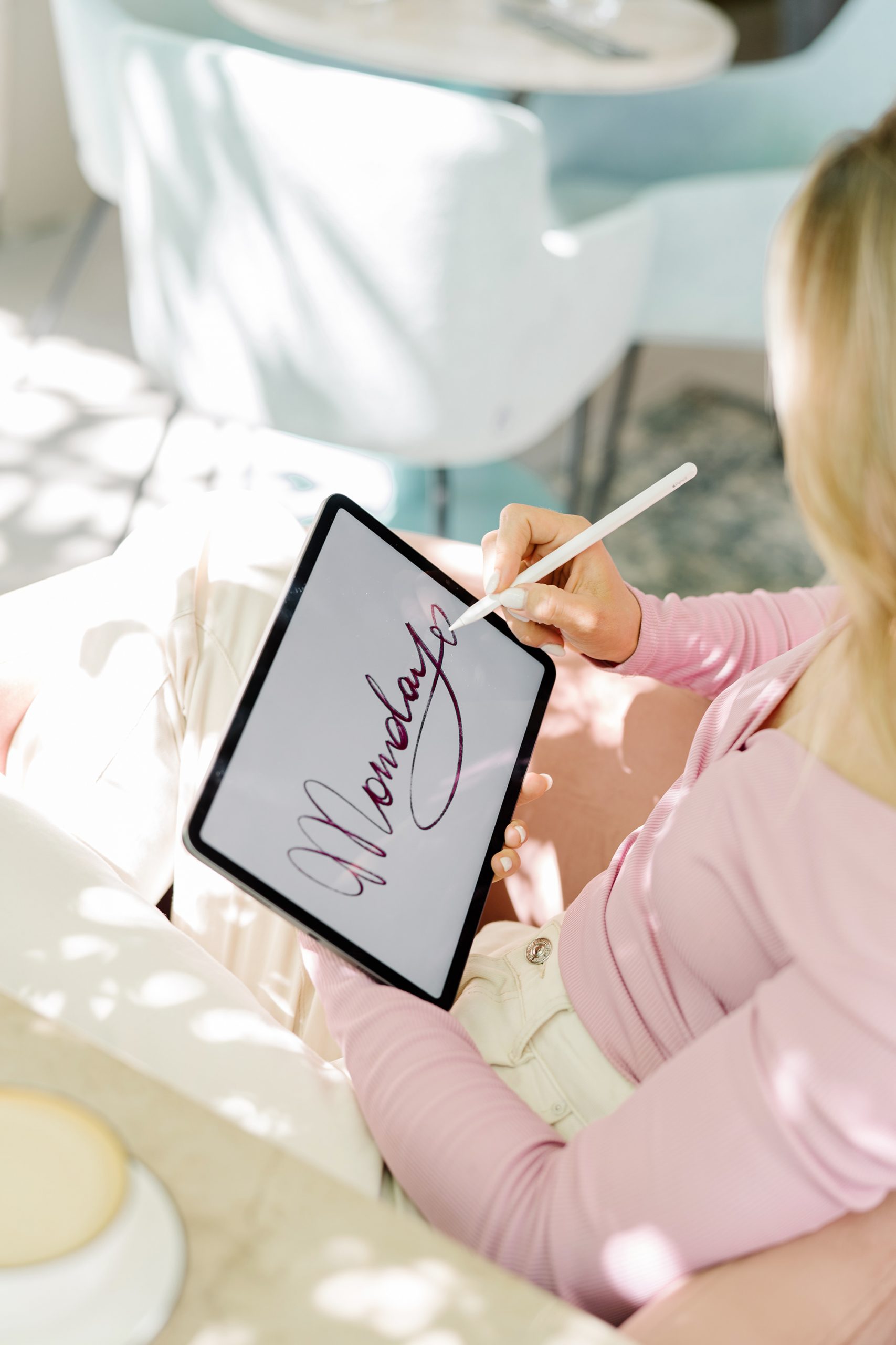 A person wearing a light pink long-sleeve top uses a stylus to write Monday in cursive on a tablet while sitting on a light-colored chair.