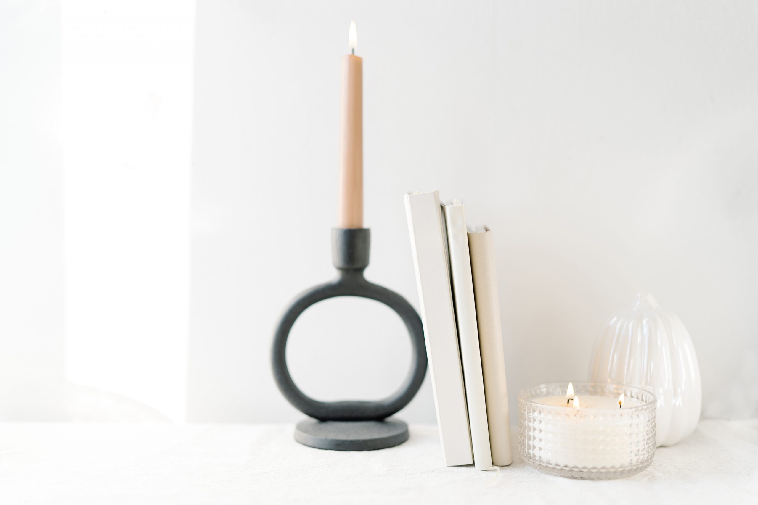 A minimalist arrangement with a lit candle in a black holder, three upright books, a small glass jar candle, and a white decorative object on a bright white surface.