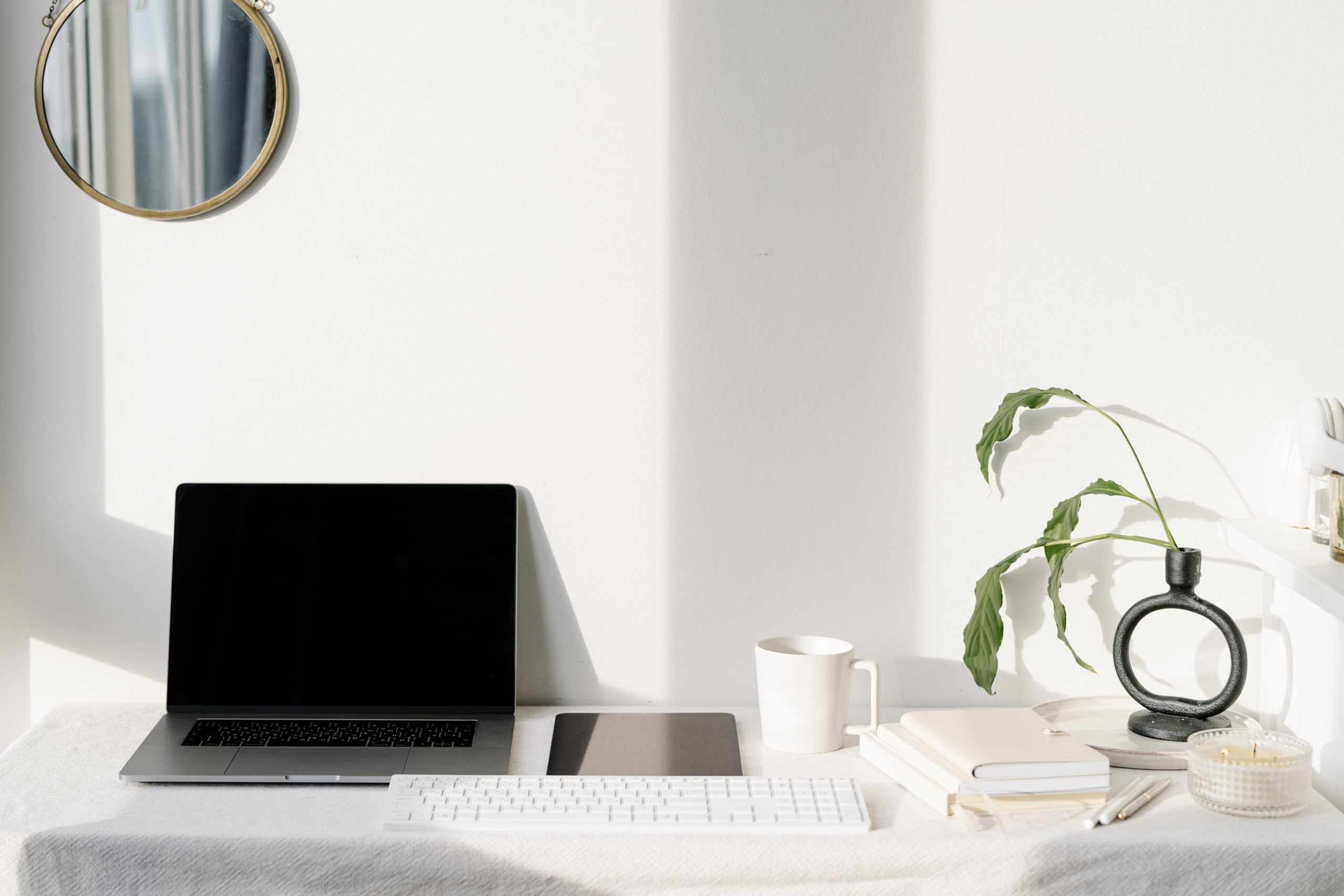 A minimalist workspace with a closed laptop, wireless keyboard, mug, plant in a round vase, notebooks, pen, and a small round mirror on a white wall, all bathed in natural sunlight.