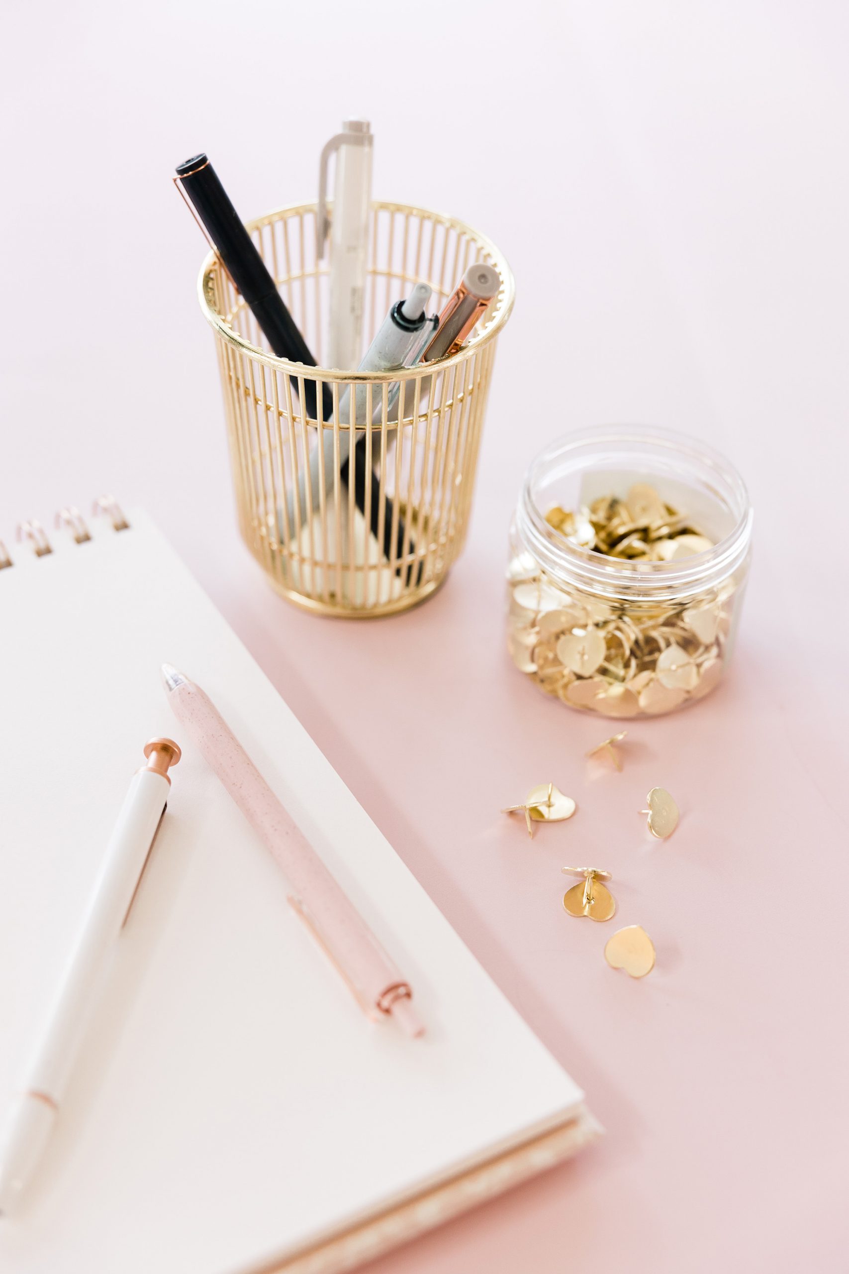 A gold pen holder with pens, a jar of gold push pins and clips, scattered pins, and a notebook with a pink and a white pen on a pastel pink surface.