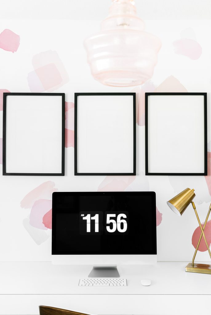 A minimalist desk setup with a computer displaying the time 11:56, a gold desk lamp, and three empty black-framed pictures on a white wall with pink and beige abstract patterns.
