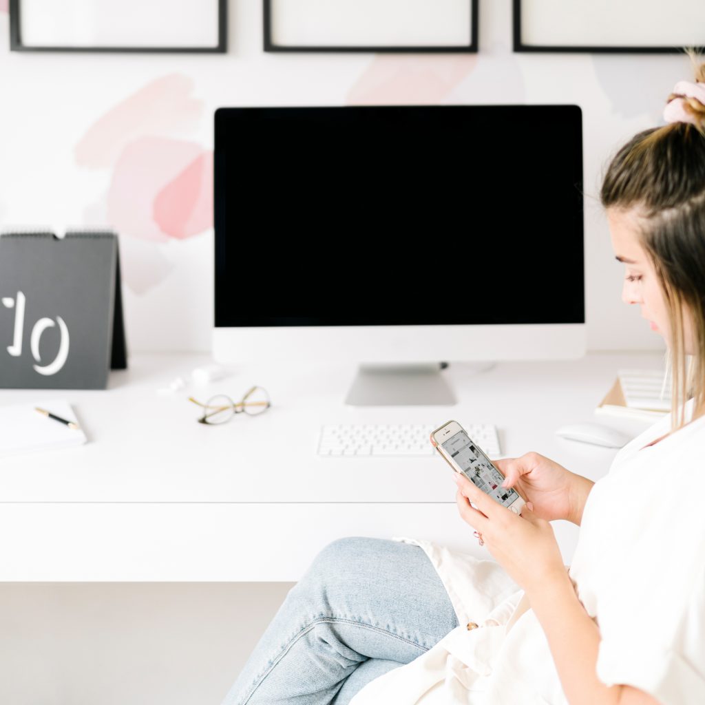A woman sits at a white desk with a computer monitor, keyboard, and calendar, looking at her smartphone. The workspace is neat and bright, with framed art and office supplies in the background.