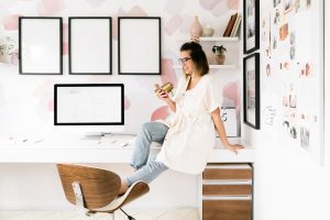 A woman in casual clothes sits on the edge of a white desk in a modern, stylish home office, holding a coffee cup and smiling. A computer, chair, and decor items are visible, with empty frames on the wall behind her.