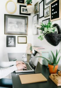 A person types on a laptop at a desk, surrounded by plants, books, and wall art in black frames. The workspace is tidy, with a closed notebook and a glass of water beside the computer.