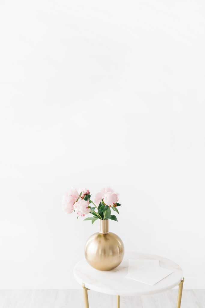 A round gold vase with light pink flowers sits on a small white table against a plain white background. An envelope or piece of paper lies on the table beside the vase.