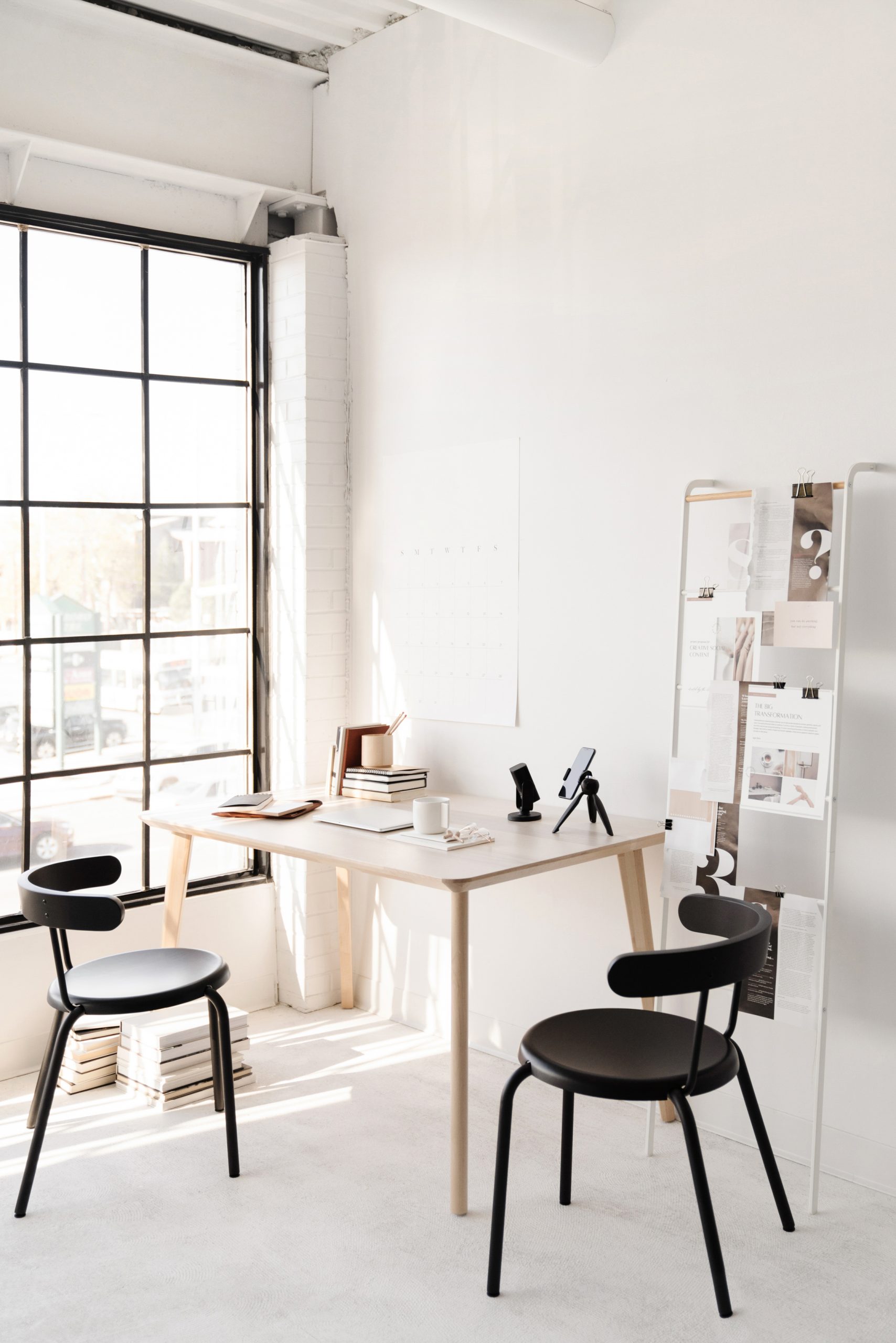A bright, minimalist office with large windows, two black chairs, a light wood table with books and stationery, and a board displaying papers and photos. Natural sunlight fills the clean, modern space.