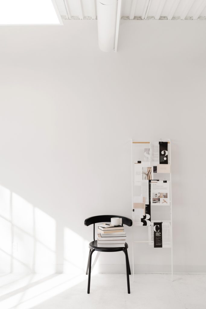 Minimalist room with white walls, a black chair stacked with books and a mug, next to a white magazine rack holding various magazines and clippings. Sunlight streams in from the left, creating soft shadows.