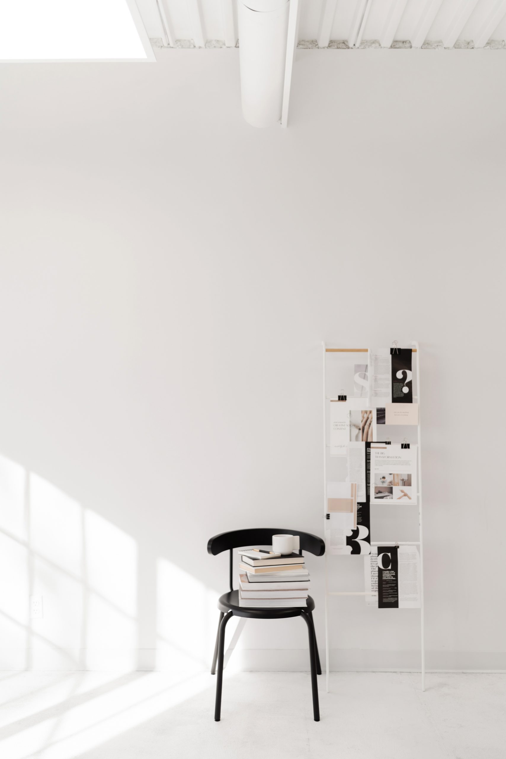 A minimalist room with white walls and floor, featuring a black chair stacked with books and a candle. Behind the chair is a magazine rack displaying various magazine pages. Sunlight streams in through a window, casting shadows.