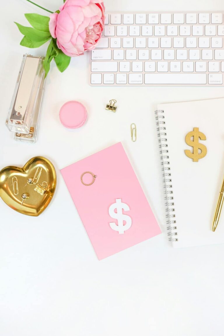 A flat lay of a white desk with a keyboard, pink flower, gold accessories, heart-shaped dish, pink notebook with a dollar sign, spiral notebook, pen, and paperclip arranged neatly.