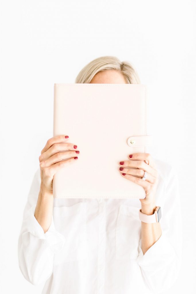 A person with light hair covers their face with a pale pink folder. They have red nail polish, wear a white shirt, a ring, and a smartwatch, standing against a white background.