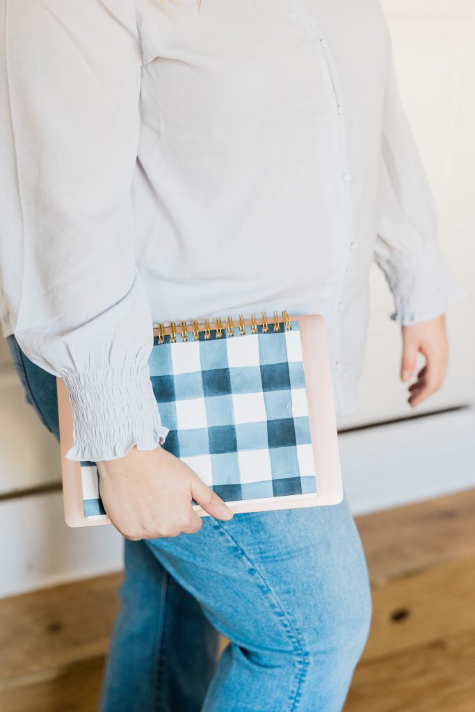A person wearing a white blouse and blue jeans holds a notebook with a blue and white checkered cover by their side. The image is cropped to show only the torso and legs.