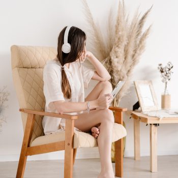 A woman with long hair wearing headphones sits on a beige chair, holding a tablet. She rests one foot on the floor and looks to the side. Dried plants and a small table decorate the bright, minimalist room.