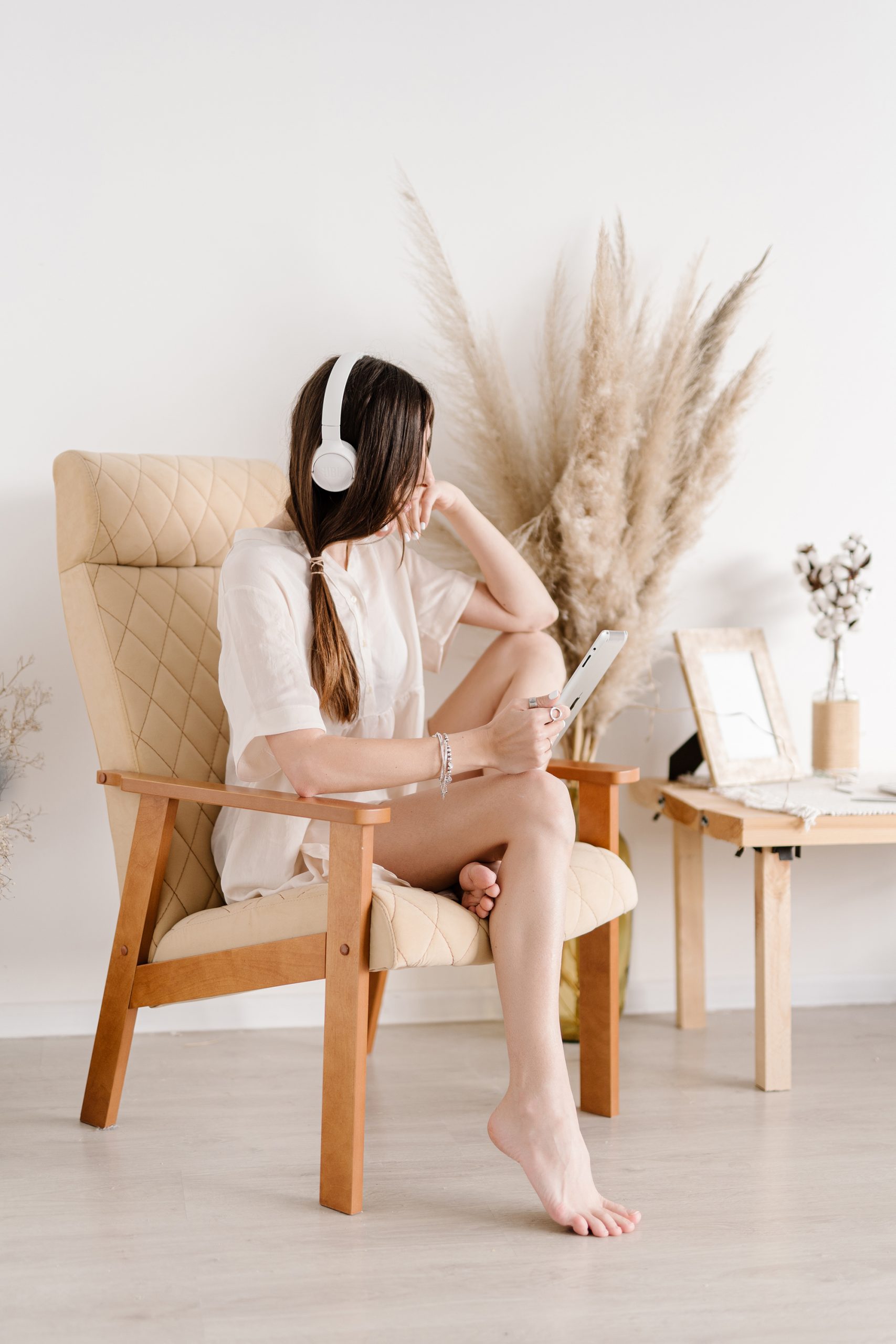 A woman with long hair wearing headphones sits on a beige chair, holding a tablet. She rests one foot on the floor and looks to the side. Dried plants and a small table decorate the bright, minimalist room.