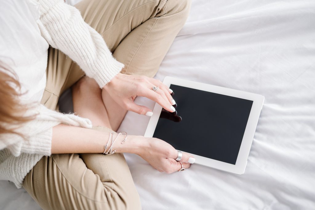 A person sitting cross-legged on a bed uses a white tablet with a blank black screen. They wear beige pants, a light sweater, and several rings and bracelets.