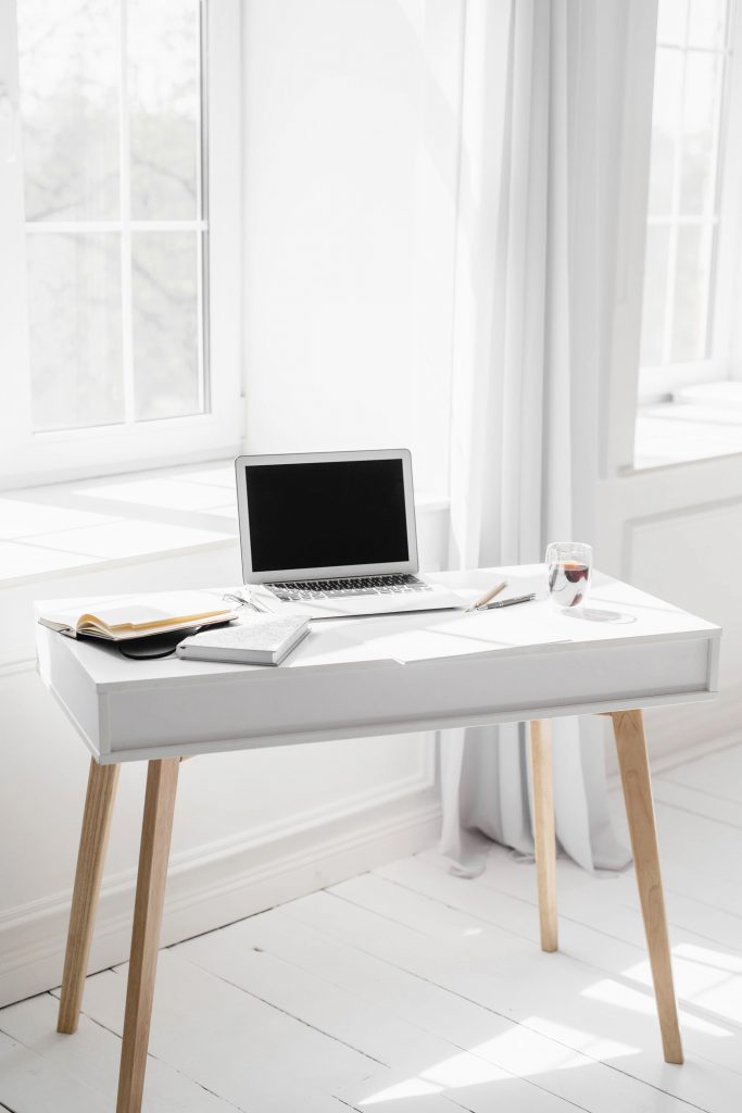 A bright, minimal workspace with a laptop, open notebook, pen, eyeglasses, and a glass of water on a white desk near a sunlit window with sheer curtains.