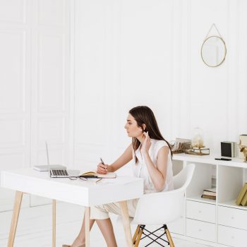 A woman sits at a white desk in a bright, minimal office, looking at her laptop while holding a pen and touching her hair. There are notebooks, books, and decor on the shelves behind her.