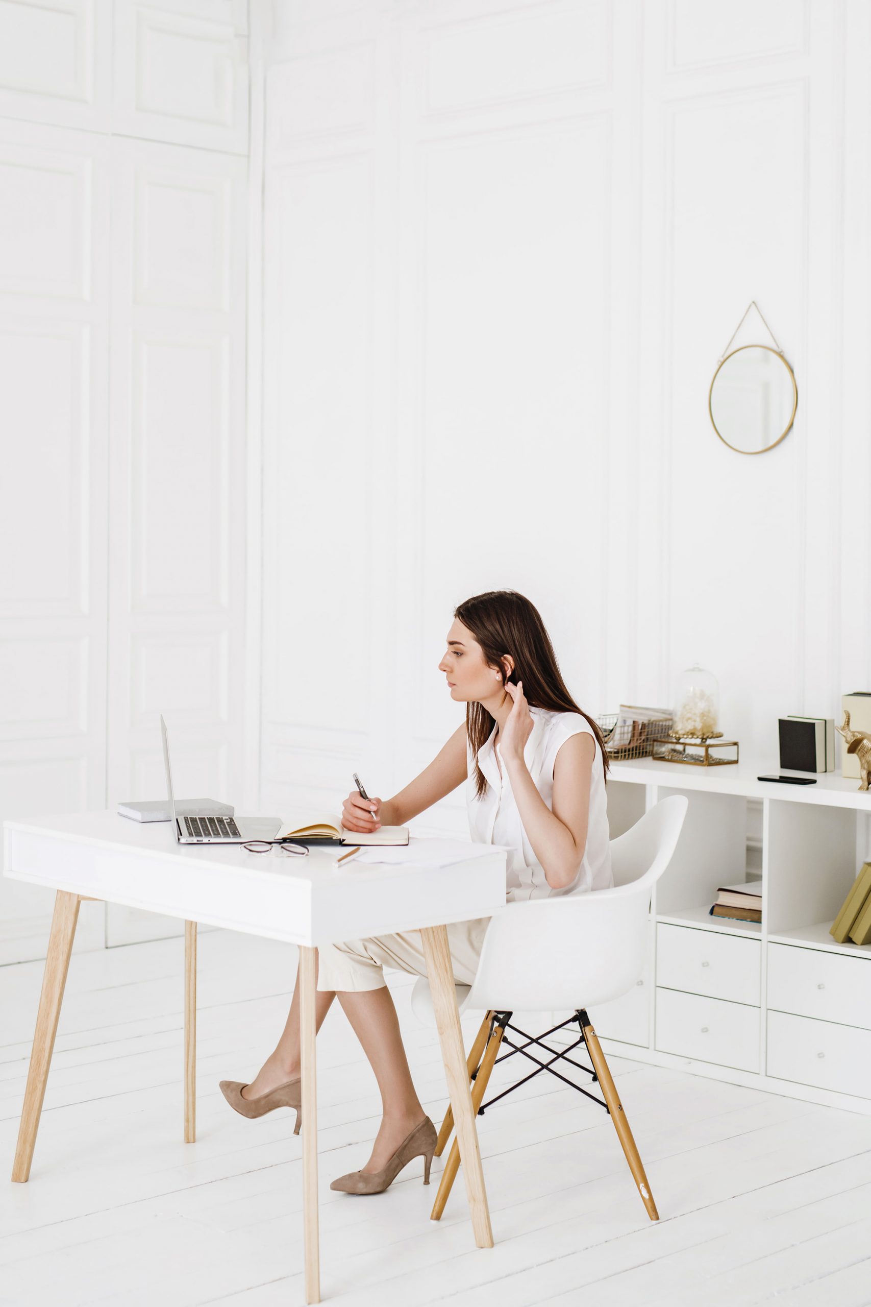 A woman sits at a white desk in a bright, minimal office, looking at her laptop while holding a pen and touching her hair. There are notebooks, books, and decor on the shelves behind her.