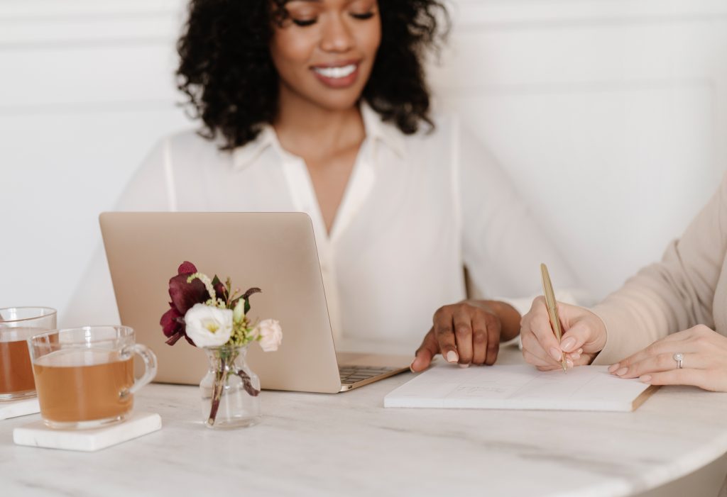 Two women sit at a table with a laptop, notepad, and tea. One is smiling and looking at the laptop, while the other writes in the notepad. A small vase with flowers is on the table.