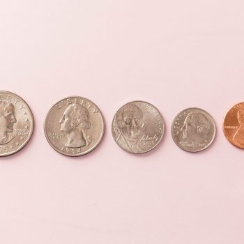 Five U.S. coins are lined up from left to right on a light pink surface: a half dollar, quarter, nickel, dime, and penny.