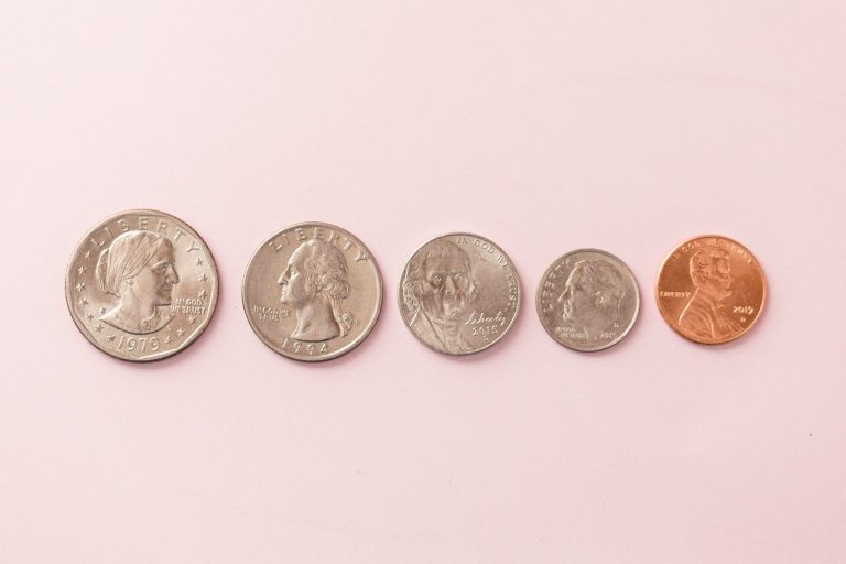 Five U.S. coins are lined up from left to right on a light pink surface: a half dollar, quarter, nickel, dime, and penny.