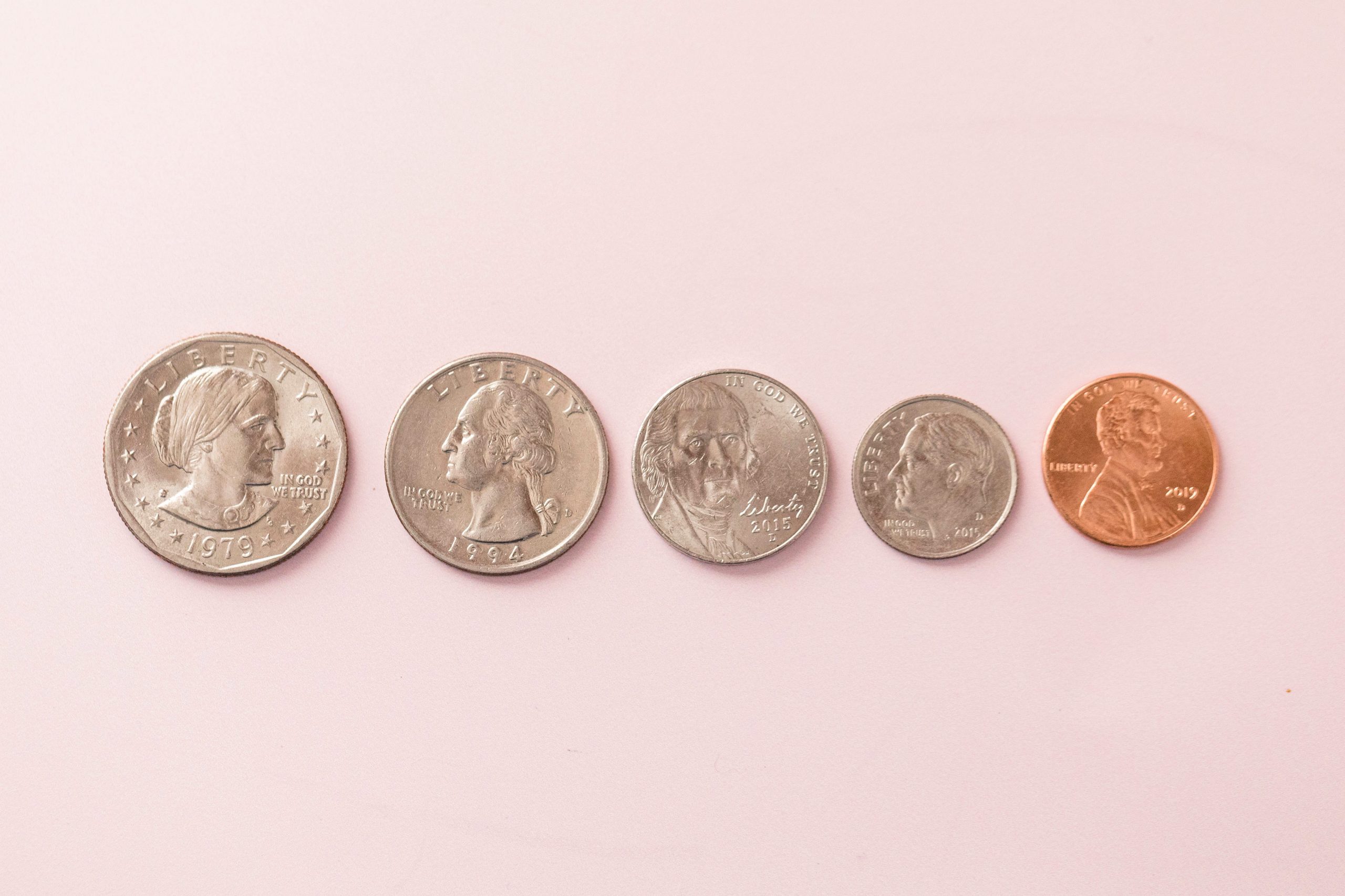 Five U.S. coins are lined up from left to right on a light pink surface: a half dollar, quarter, nickel, dime, and penny.