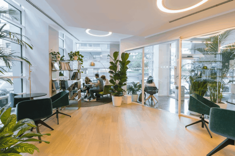 Modern office lounge with green chairs, potted plants, bookshelves, and wooden floors. A group of people are having a meeting at a table near large windows with natural light streaming in.