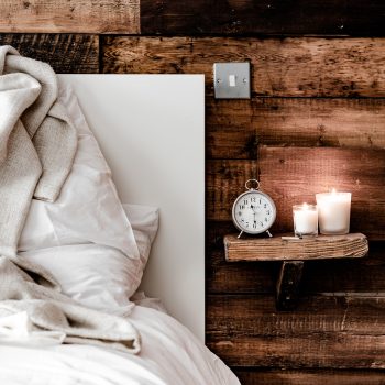 A cozy bed with white linens and a beige blanket sits next to a rustic wooden wall. A small shelf holds a silver alarm clock and two lit candles, creating a warm, inviting atmosphere.