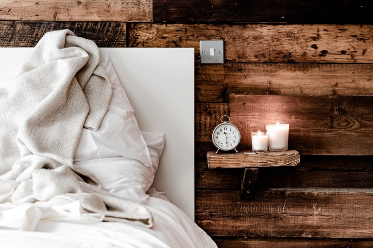 A cozy bed with white linens and a beige blanket sits next to a rustic wooden wall. A small shelf holds a silver alarm clock and two lit candles, creating a warm, inviting atmosphere.
