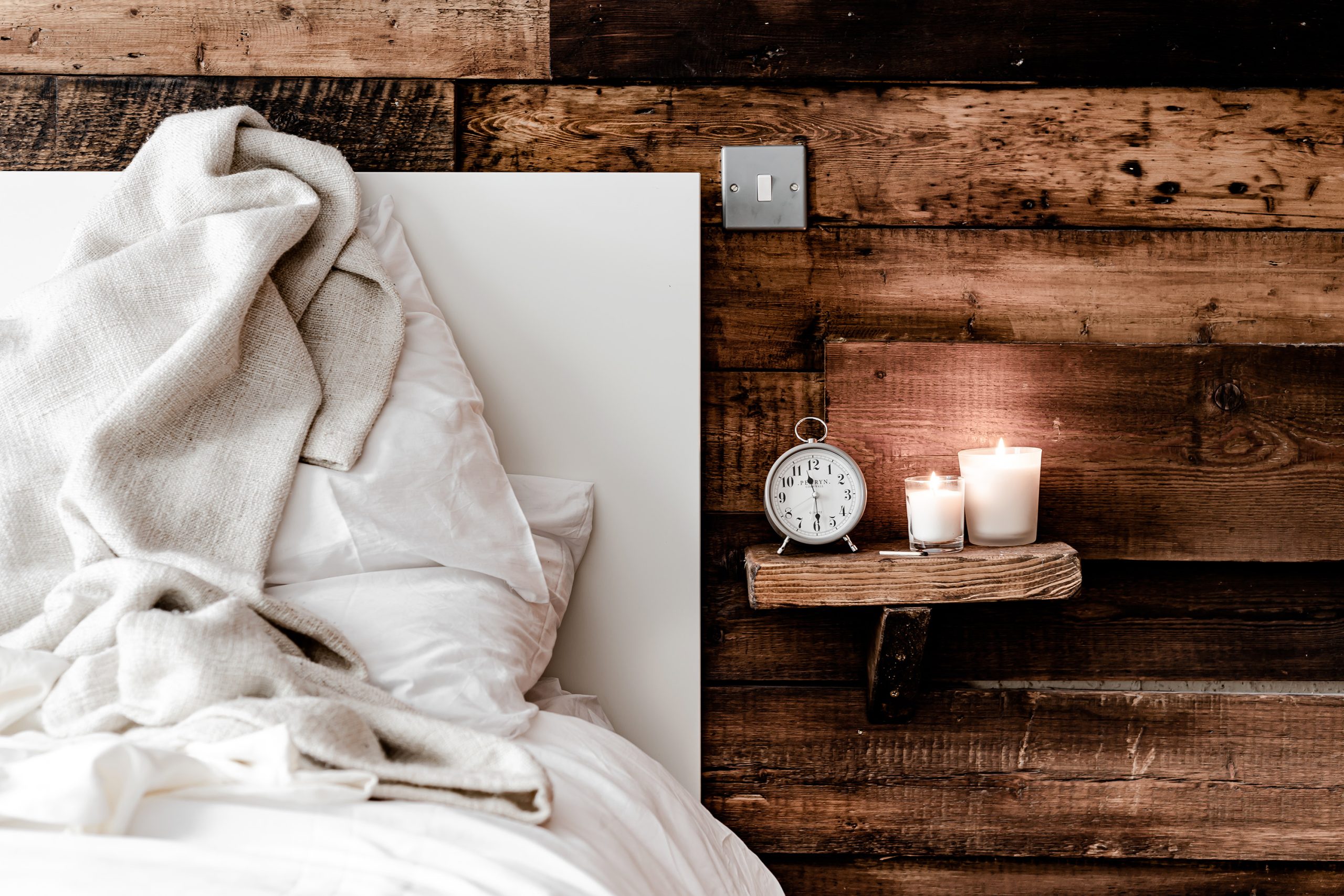A cozy bed with white linens and a beige blanket sits next to a rustic wooden wall. A small shelf holds a silver alarm clock and two lit candles, creating a warm, inviting atmosphere.